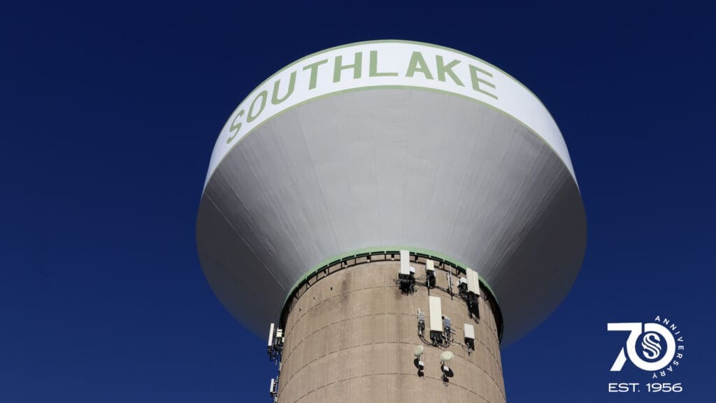 A tall water tower with "SOUTHLAKE" written at the top, set against a clear blue sky. The bottom right corner features a 70th anniversary logo and "EST. 1956.
