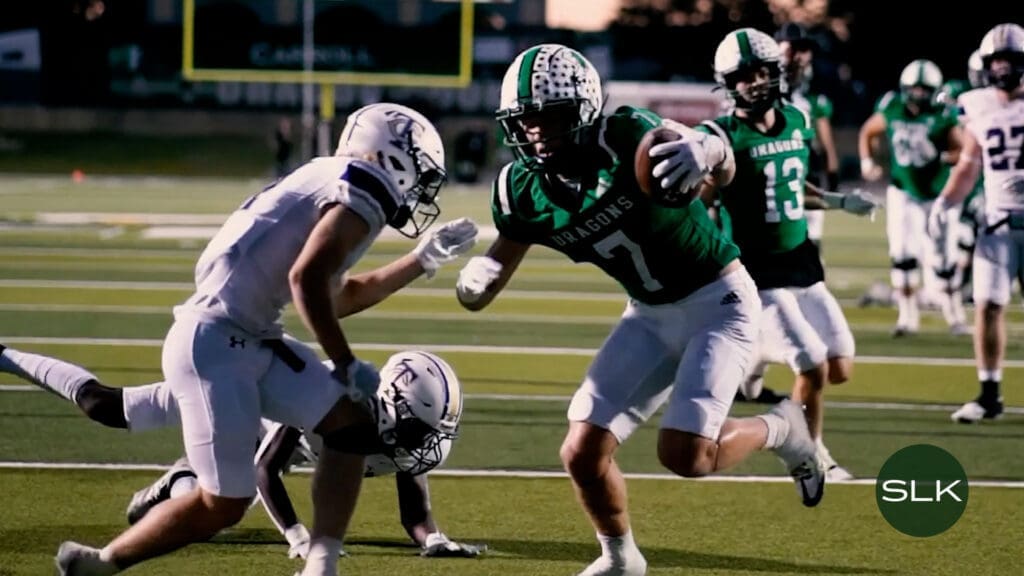 A football player in a green "Dragons" jersey runs with the ball, dodging two defenders in white uniforms on a field. Other players and a goalpost are visible in the background. An "SLK" icon is seen in the bottom right corner.