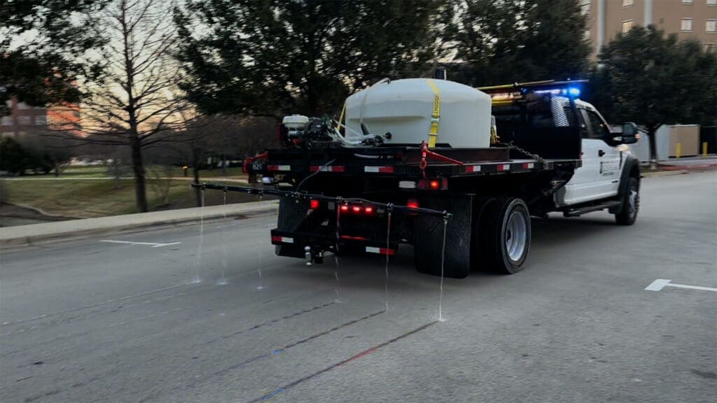 A truck with a mounted tank sprays liquid onto the road from multiple nozzles at the rear, likely for de-icing or road treatment, driving on a city street near trees and buildings.