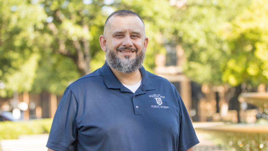 A man with a beard smiles outdoors, wearing a navy blue "Southlake Public Works" polo shirt. Trees and a blurred fountain are visible in the sunny background.