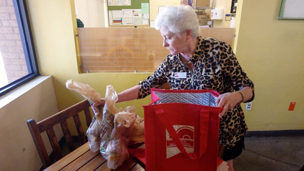 An elderly woman wearing a leopard-print shirt unpacks groceries from plastic bags into a red reusable tote bag on a wooden table in a brightly lit room.