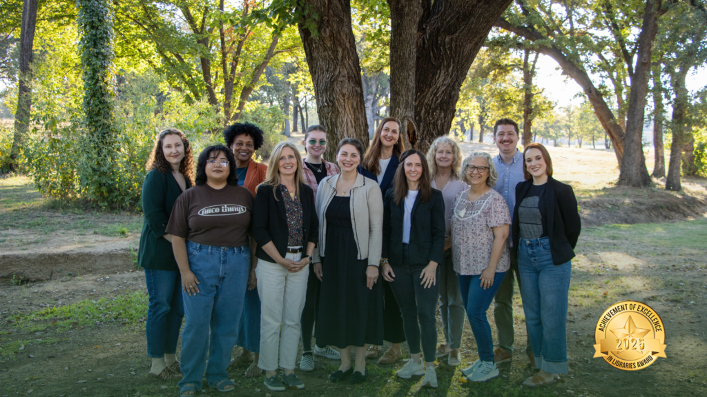 A diverse group of twelve adults stands together smiling in a sunny park with tall trees and green grass. A gold “Achievement of Excellence 2025” badge appears in the lower right corner of the image.