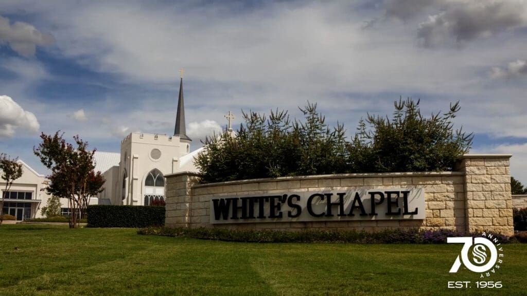 A large stone sign reading "WHITE'S CHAPEL" stands in front of a church with a tall steeple. The lawn is green, and the sky is partly cloudy. A “70th Anniversary EST. 1956” logo appears in the corner.