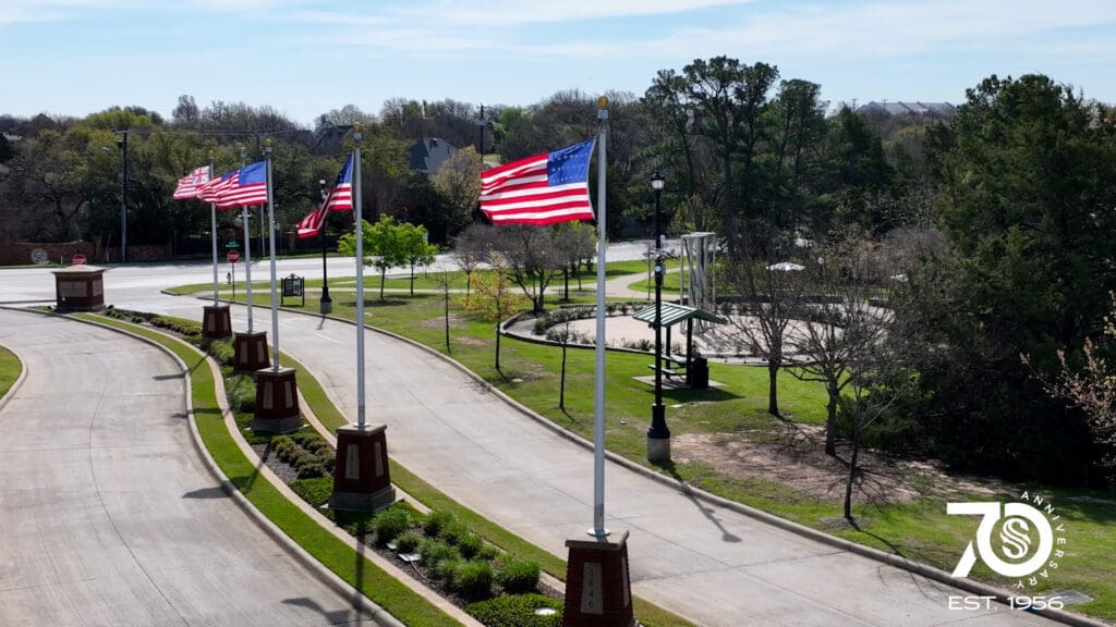 Five American flags on poles line a curved road with grass, trees, and a small pavilion nearby. A "70th Anniversary EST. 1956" logo appears in the lower right corner. The sky is clear and sunny.