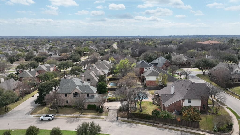 A suburban neighborhood with houses, trees, and winding streets under a partly cloudy sky. Some trees have sparse leaves, indicating early spring or late fall. A white car is parked on the street in the foreground.