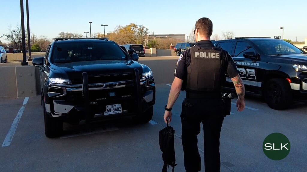 A police officer in uniform faces two parked police SUVs in a parking lot, holding a bag in one hand. The vehicles display "POLICE" markings and have Texas license plates. Trees and buildings are in the background.