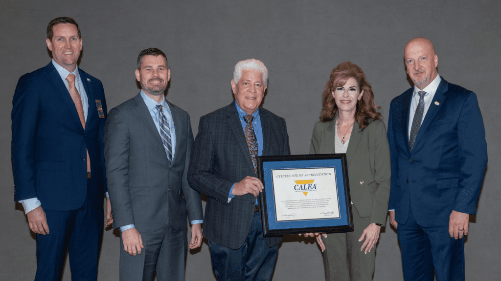 Five people in business attire stand together. The person in the center holds a framed "Certificate of Accreditation" from CALEA. They all smile and pose against a plain gray background.