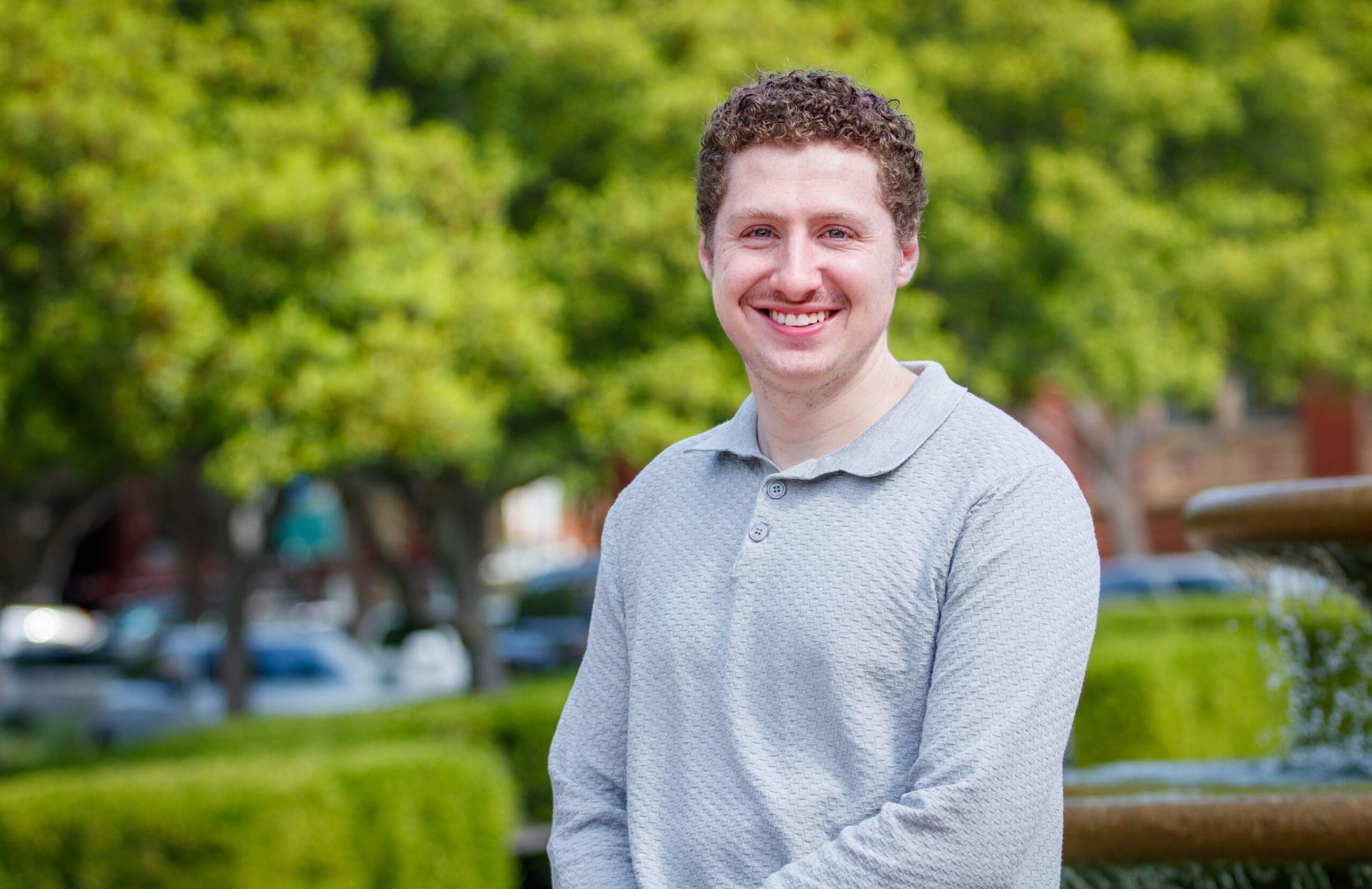 A man with short curly hair, wearing a light gray long-sleeve shirt, smiles while standing outdoors in front of green trees and a fountain.