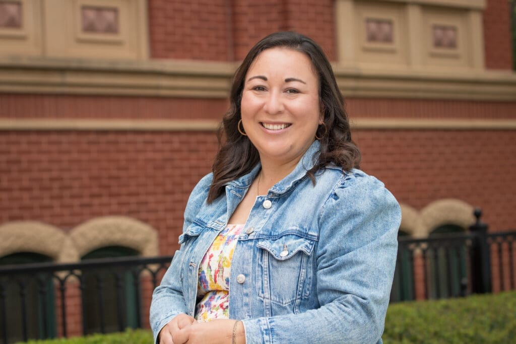 A woman with long brown hair, wearing a denim jacket and a colorful top, smiles while standing outdoors in front of a red brick building with green-windowed arches and trimmed greenery.