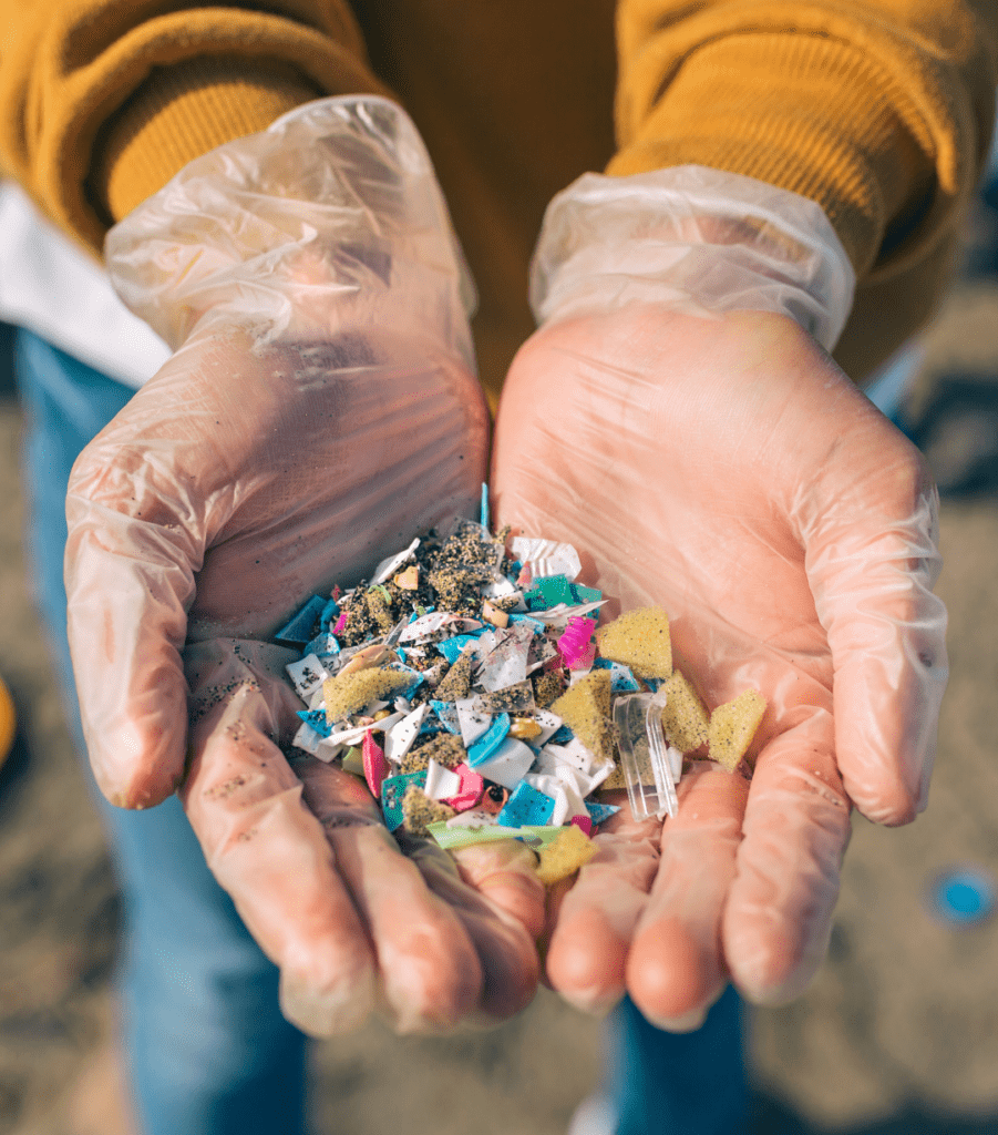 A person wearing disposable gloves holds a handful of colorful plastic fragments and debris, likely collected from a beach, highlighting pollution and environmental concerns.
