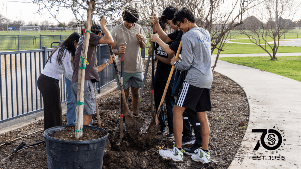 A group of young people and an adult work together to plant a tree in a park, using shovels. The scene is outdoors on a sunny day, and a 70th anniversary logo appears in the corner.