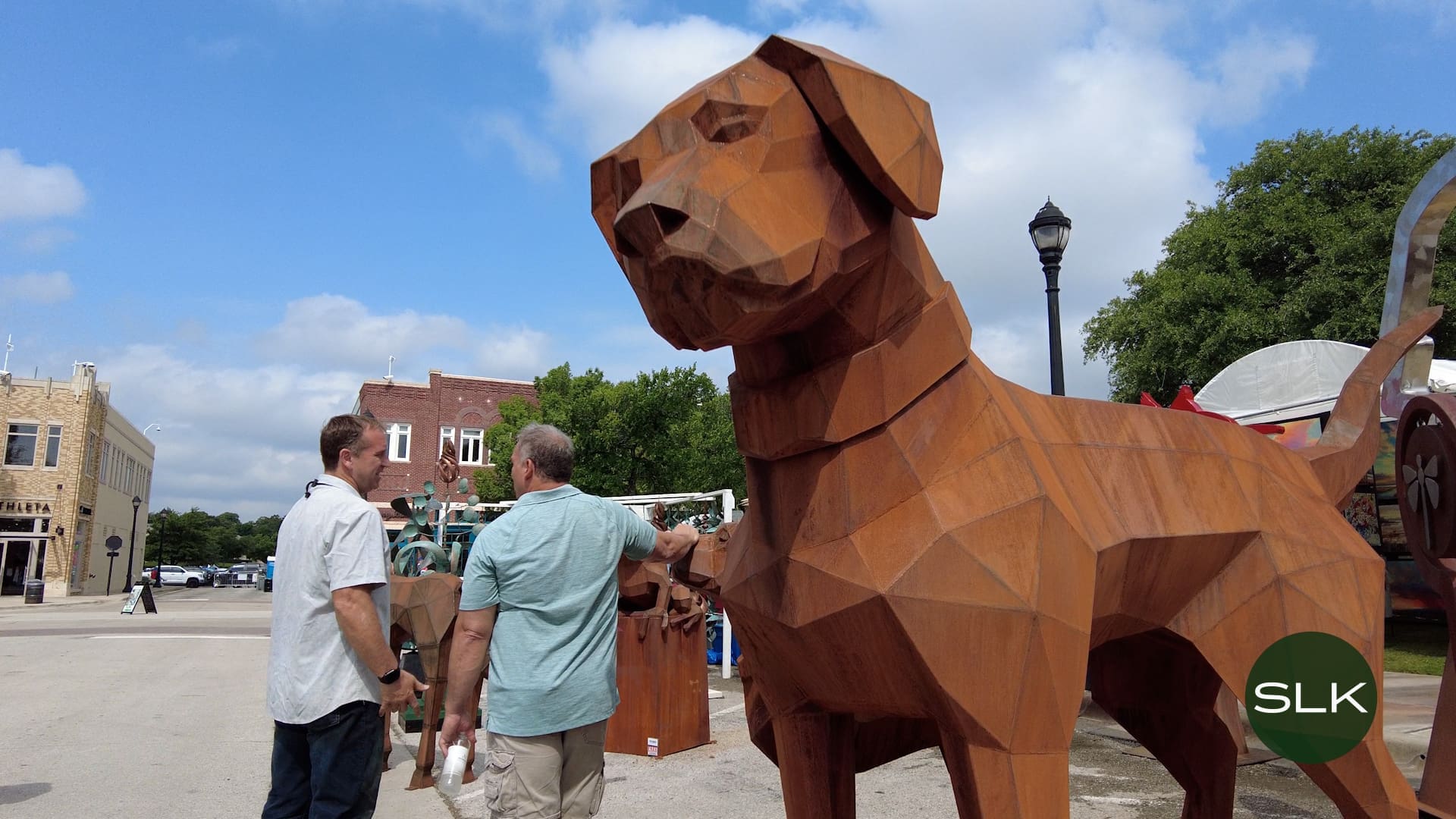 Two men stand next to a large, angular wooden dog sculpture in a town square with brick buildings, trees, and a streetlamp visible in the background.