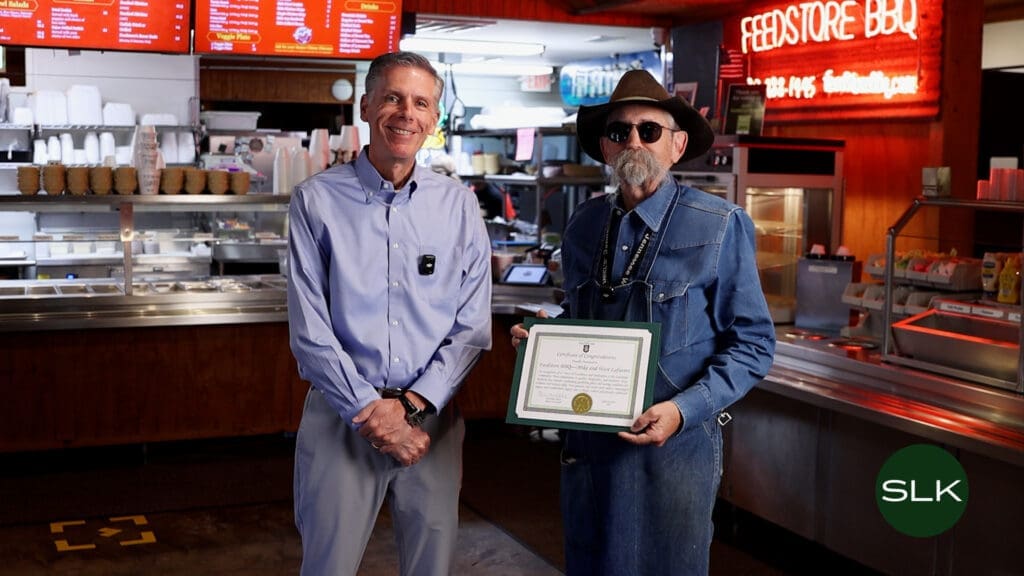 Mayor Shawn McCaskill and Mike Lafavers stand inside a BBQ restaurant; one is dressed casually and smiling, while the other wears sunglasses, a hat, and denim, holding a framed certificate. The counter and menu boards are visible in the background.