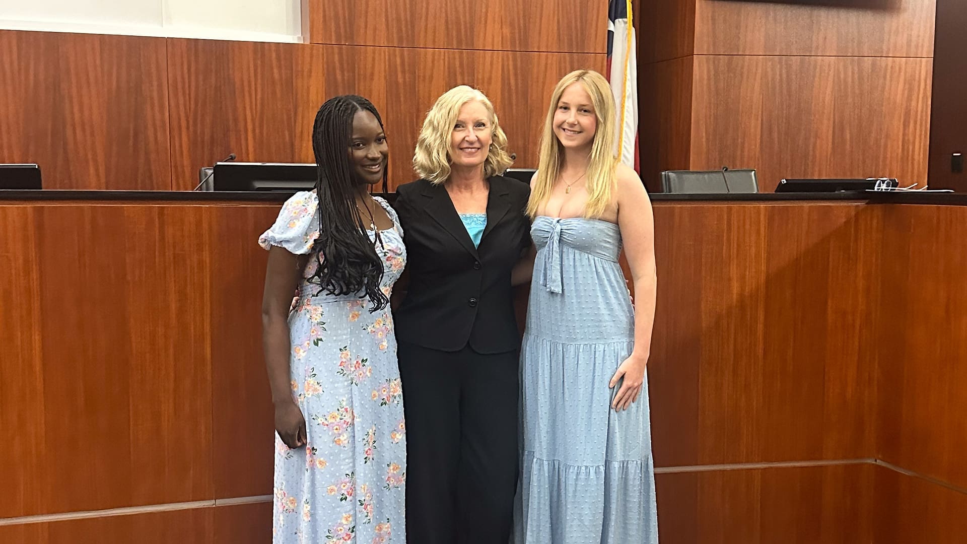 Three women stand together in a courtroom with wooden walls. Two women wear light blue dresses with floral patterns, and the woman in the middle wears a black suit. They are smiling and standing in front of a judge’s bench and an American flag.