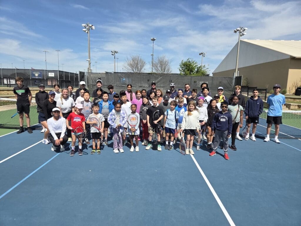 A large group of children and adults pose together on an outdoor tennis court, smiling for the camera. Most are holding tennis rackets, and the weather appears sunny with a clear sky.