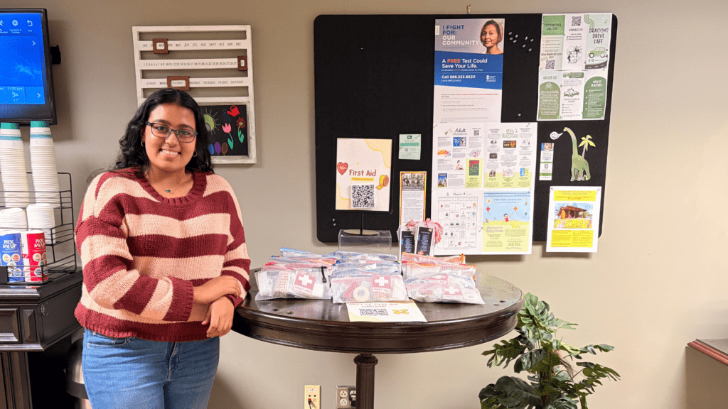 A woman wearing glasses and a striped sweater stands by a round table with several first aid kits in plastic bags. Behind her, a bulletin board displays various community flyers and posters.