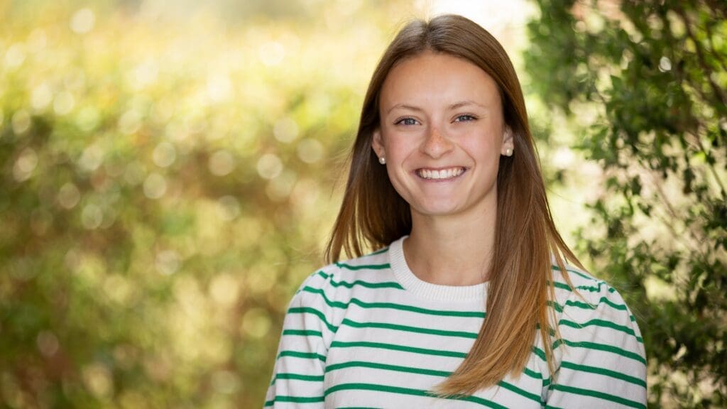 A young woman with straight light brown hair, wearing a white and green striped shirt, smiles while standing outdoors in front of blurred greenery.