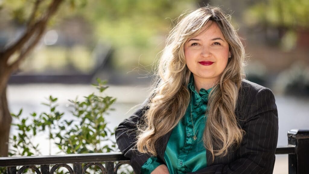 A woman with long blonde hair and red lipstick, wearing a teal blouse and dark pinstripe blazer, stands outdoors near a metal railing with greenery and sunlight in the background.