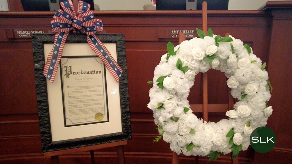 A framed proclamation with a patriotic ribbon stands next to a large white flower wreath on a wooden easel in a council chamber; wooden benches and nameplates are visible in the background.