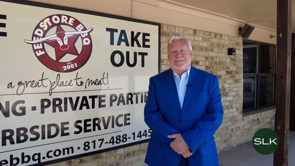Councilmember Gary Fawks smiles as he wears a blue suit and stands in front of a Feedstore BBQ restaurant sign on a brick wall.