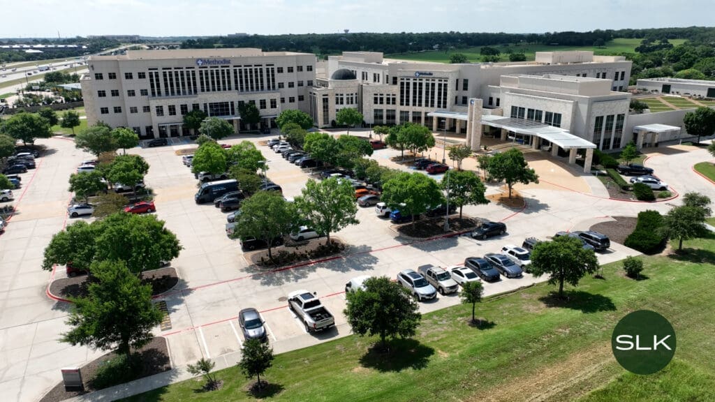 Aerial view of a large medical office building with a parking lot filled with cars, surrounded by green trees and grass under a partly cloudy sky. The building is labeled "Methodist Southlake Medical Center" A green circular icon in the bottom right corner says "SLK".