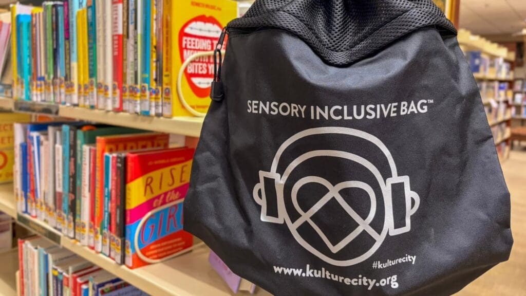 A black "Sensory Inclusive Bag" hangs on a library bookshelf filled with colorful books. The bag displays a logo with headphones and a heart, along with the website www.kulturecity.org.