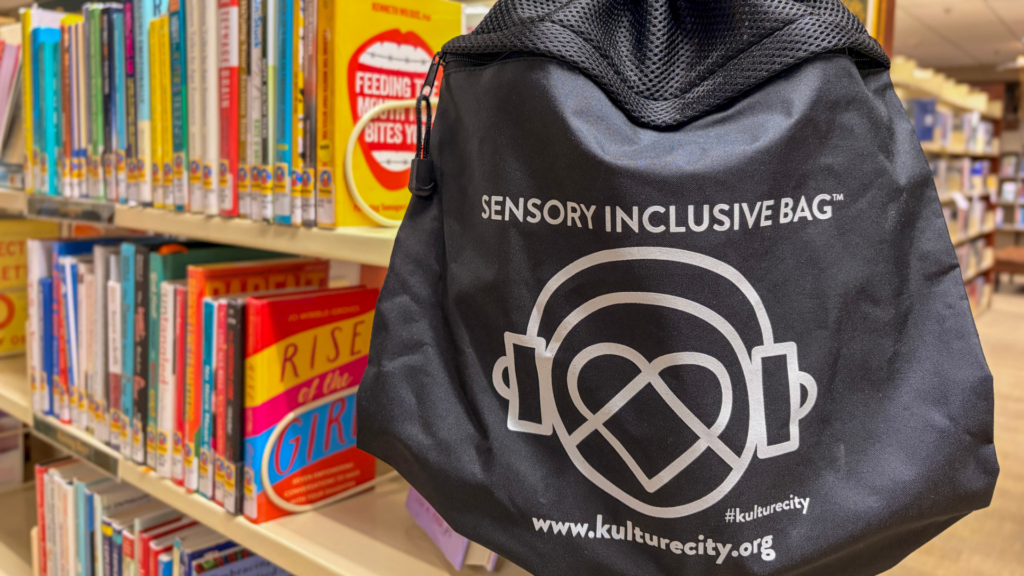 A black sensory inclusive bag with a headphone and heart logo hangs on a library shelf filled with colorful books. The bag displays text and a website, kulturcity.org, promoting sensory accessibility.