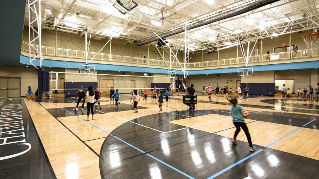 Children and adults play volleyball and basketball in a large, well-lit indoor gymnasium with high ceilings, multiple courts, and a running track above. The gym is busy with activity and groups engaging in different sports.