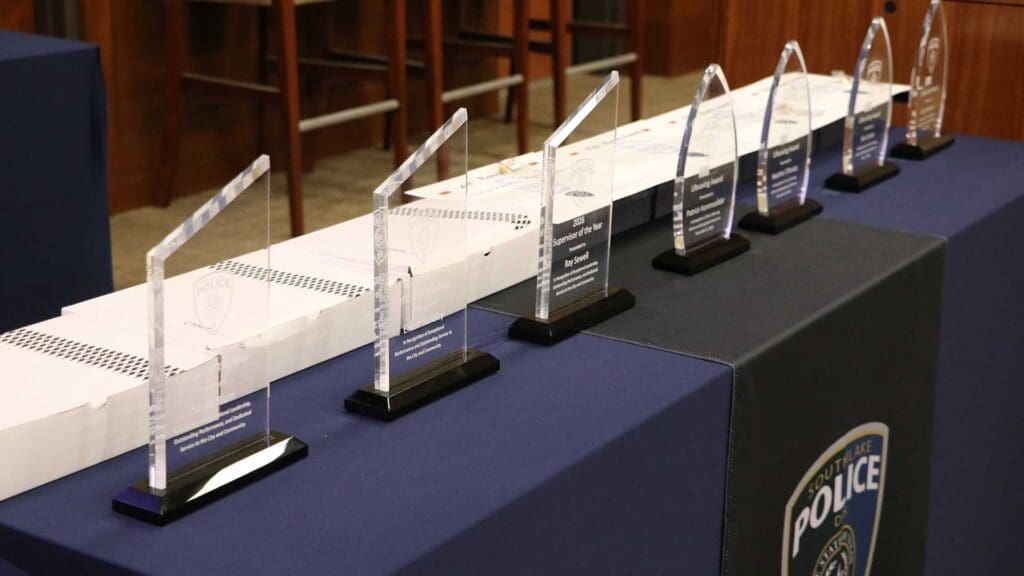 A row of clear glass police awards with black bases is displayed on a blue tablecloth, with the edge of a police department seal visible and boxes in the background.