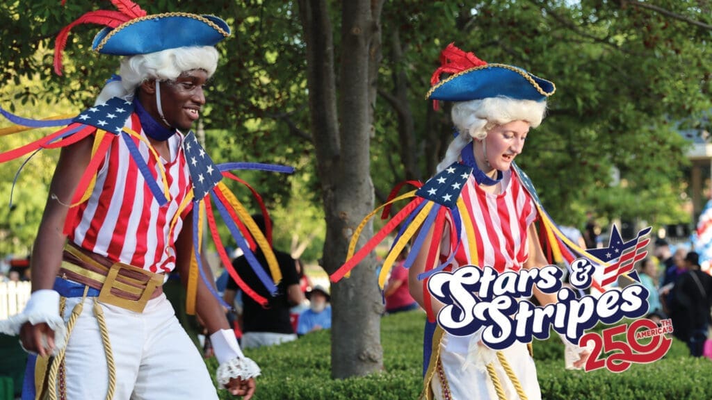 Two people in colorful colonial-style costumes with American flag patterns and feathered hats walk outdoors. A "Stars & Stripes 250" logo is in the corner, celebrating America's 250th anniversary. Trees and people are in the background.