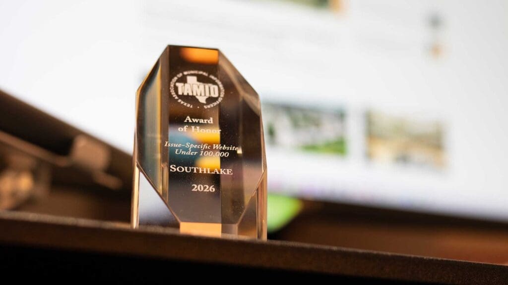 A close-up of a crystal trophy reading "TAMIO Award of Honor, Issue-Specific Website Under 100,000, Southlake, 2026," sitting on a dark surface with a blurred computer screen in the background.