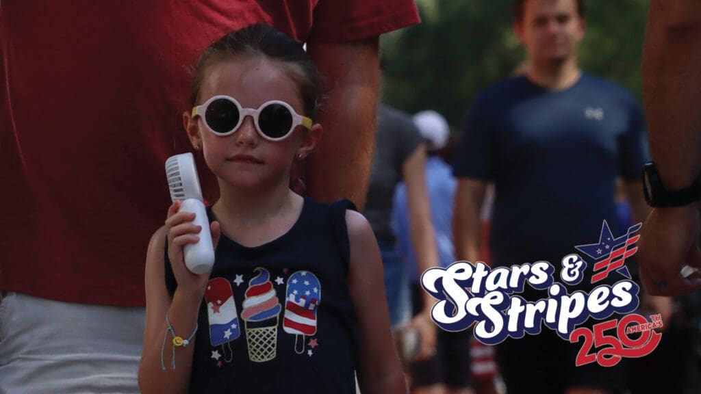 A young girl wearing round white sunglasses and a navy blue shirt with patriotic popsicle designs holds a white object, standing among people at an outdoor event. The "Stars & Stripes 250" event logo appears in the corner.