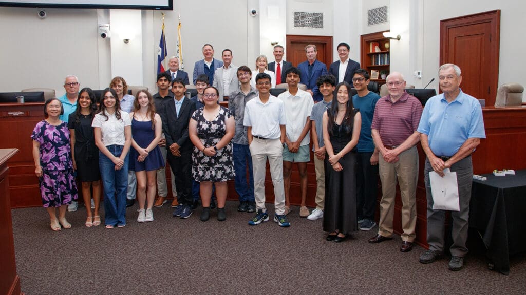 A diverse group of students and adults stand together in a courtroom or council chamber, posing for a group photo in front of wooden desks, a Texas flag, and two American flags.