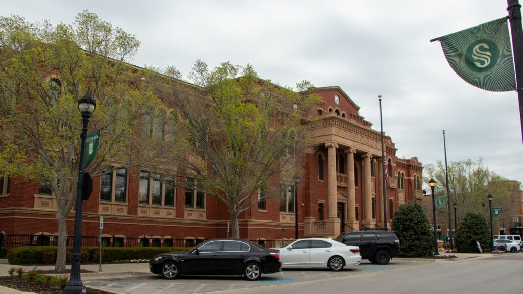 A large red-brick building with tall windows and columns, partly obscured by trees. Cars are parked in front, and green street banners with an “S” logo hang from poles. The sky is overcast.