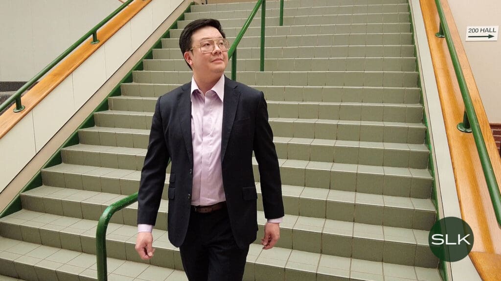 Councilmember Eric Wang, wearing a suit and glasses, walks down a tiled staircase with green railings inside a building. There is a sign on the wall indicating "200 Hall" and a circular logo with "SLK" in the bottom right corner.
