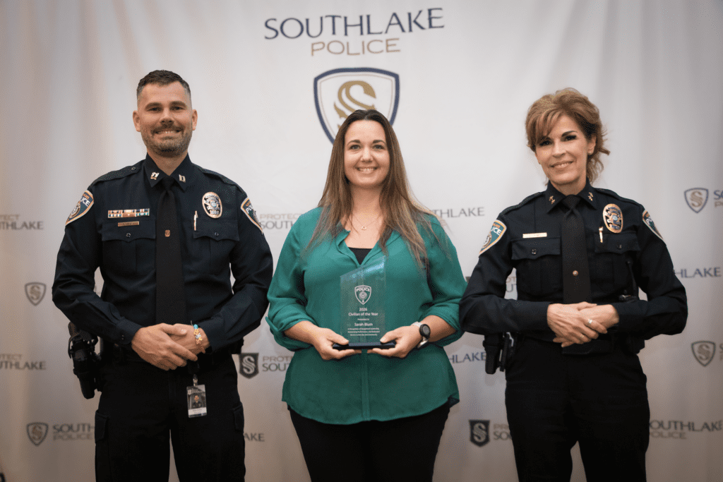 Three people pose in front of a Southlake Police backdrop. The woman in the middle holds an award, flanked by two uniformed police officers smiling at the camera.