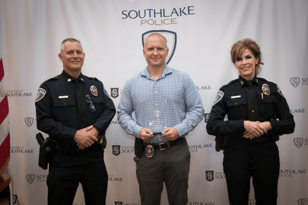 Three people stand in front of a Southlake Police backdrop, two in police uniforms on either side of a man in a blue checkered shirt holding an award. All are smiling.