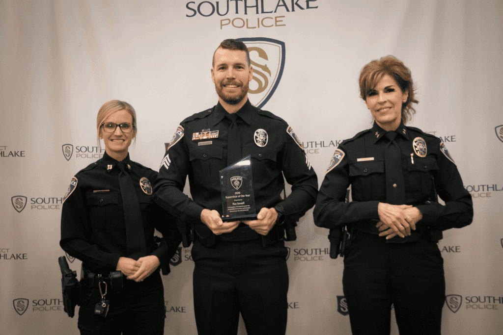 Three uniformed police officers stand in front of a "Southlake Police" banner. The officer in the center holds an award plaque and all three are smiling at the camera.