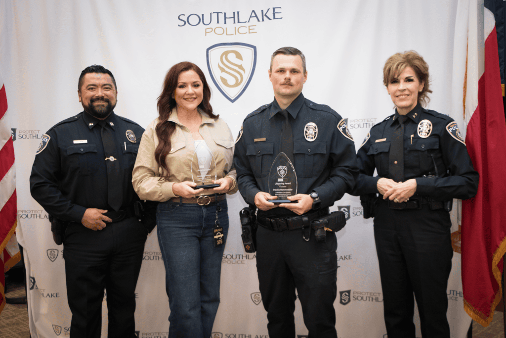 Four people stand in front of a Southlake Police backdrop. Two police officers in uniform stand on either end, while a woman in a tan blouse and a man in uniform hold awards in the center.
