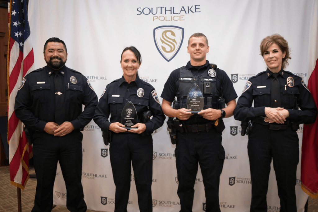 Four Southlake Police officers stand in uniform, three holding awards, posing and smiling in front of a Southlake Police backdrop with American flags at the sides.
