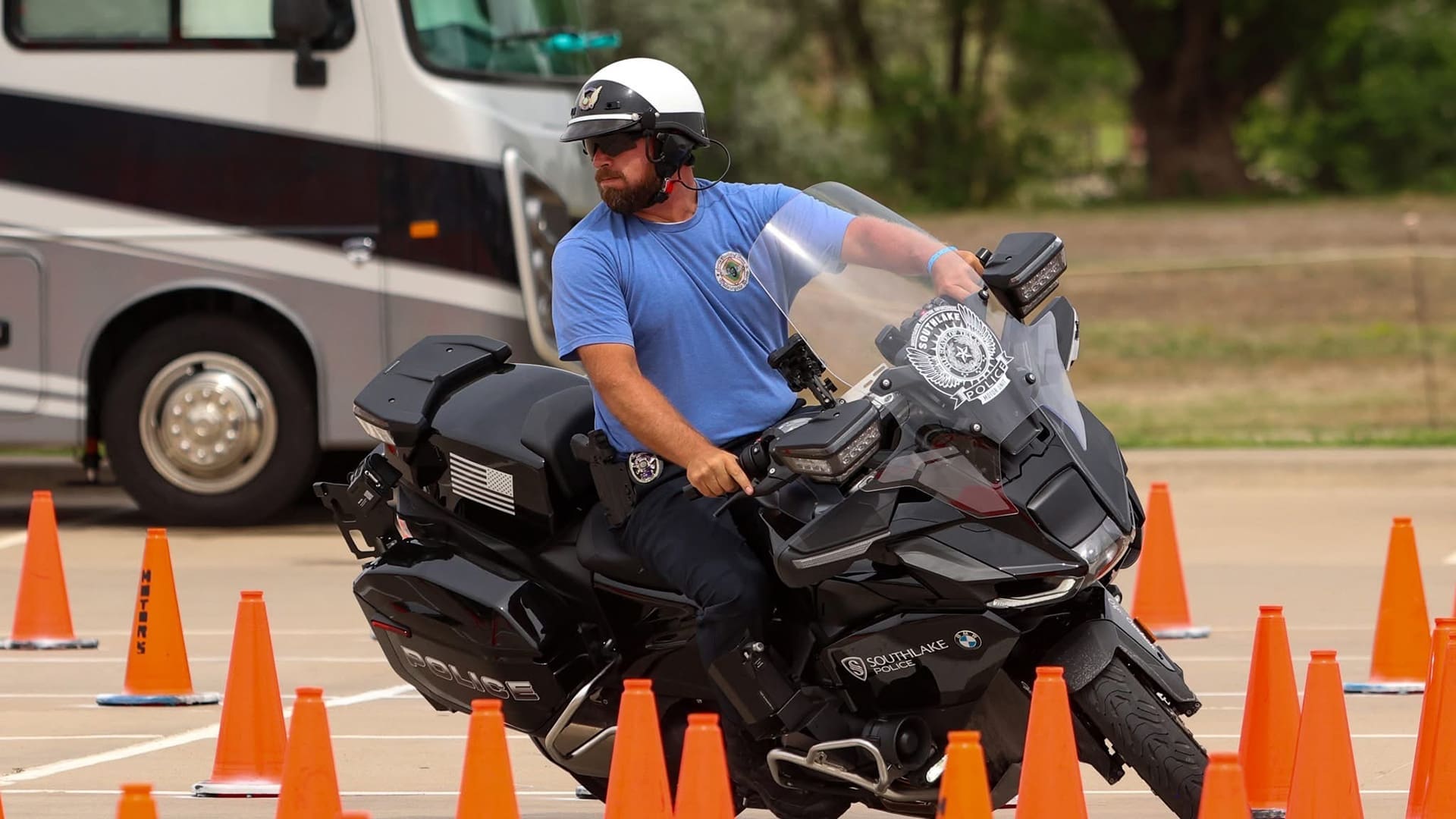 A police officer in a blue shirt and helmet rides a black motorcycle through an obstacle course marked with orange traffic cones, with a bus parked in the background.