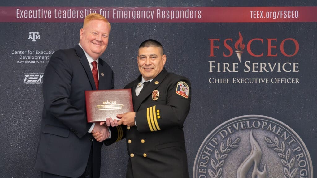 Two men stand smiling and shaking hands in front of a backdrop for Fire Service Chief Executive Officer training. One man wears a suit, the other a fire chief uniform, holding a certificate.
