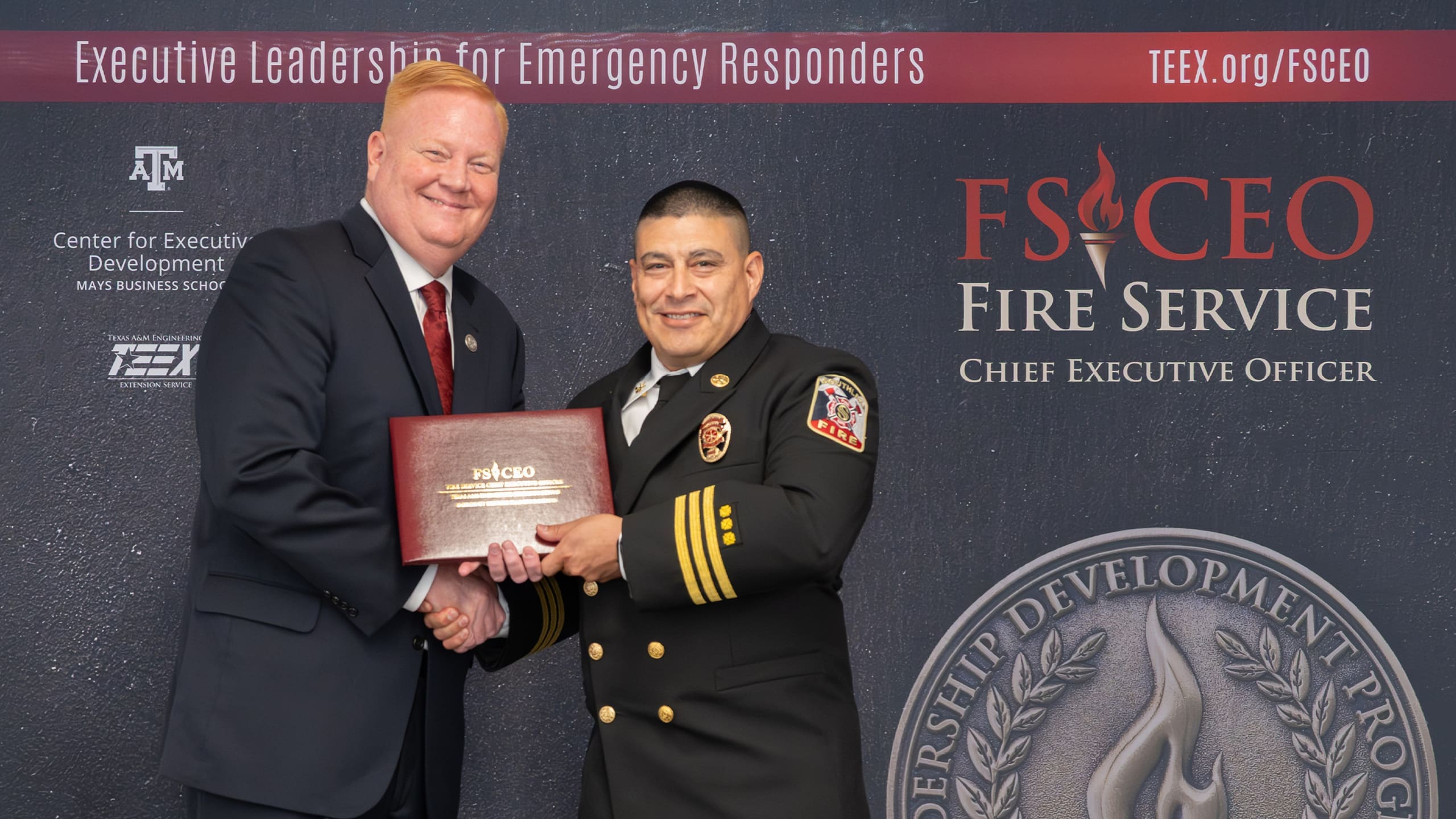 Two men stand smiling and shaking hands in front of a backdrop for Fire Service Chief Executive Officer training. One man wears a suit, the other a fire chief uniform, holding a certificate.
