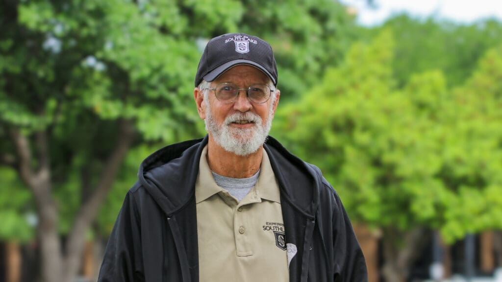 An older man with a gray beard, glasses, and a dark cap, wearing a black jacket over a beige polo shirt, stands outdoors in front of green trees, smiling at the camera.