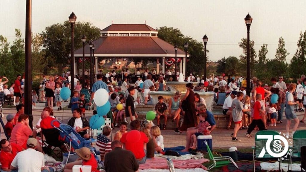 A crowd gathers in a park by a gazebo, many holding balloons and sitting on blankets. Children and adults enjoy the outdoor event. A "70" anniversary logo is in the lower right corner.