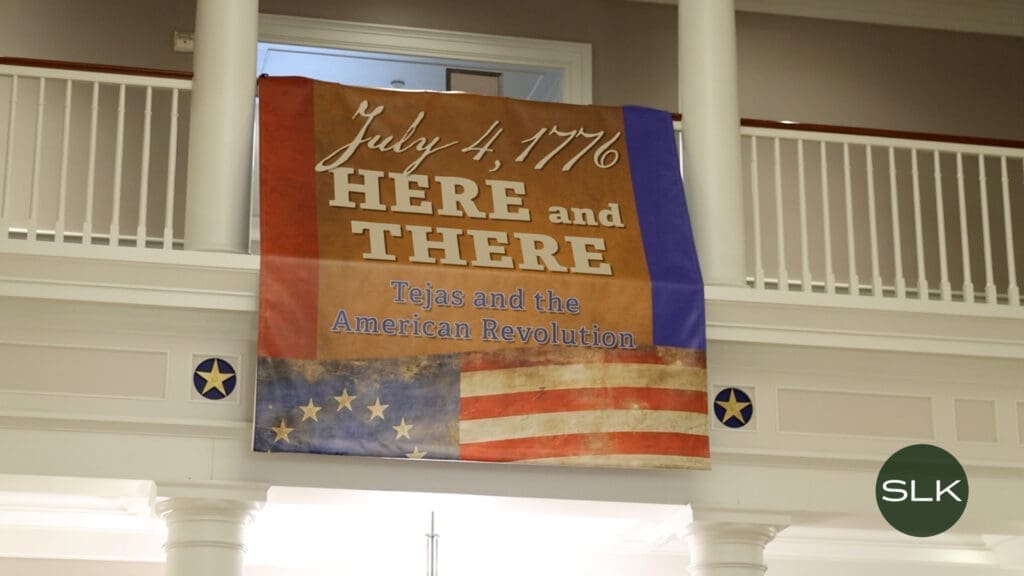 A banner hangs on a balcony, reading "July 4, 1776 HERE and THERE Tejas and the American Revolution," with images of a colonial American flag and the Texas flag. White columns and stars decorate the balcony.
