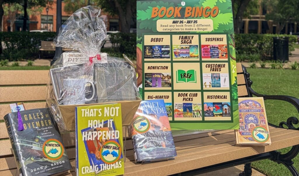 A park bench displays a Book Bingo board, four books, and a gift basket with a mug and snacks. A grassy park and trees are visible in the sunny background.